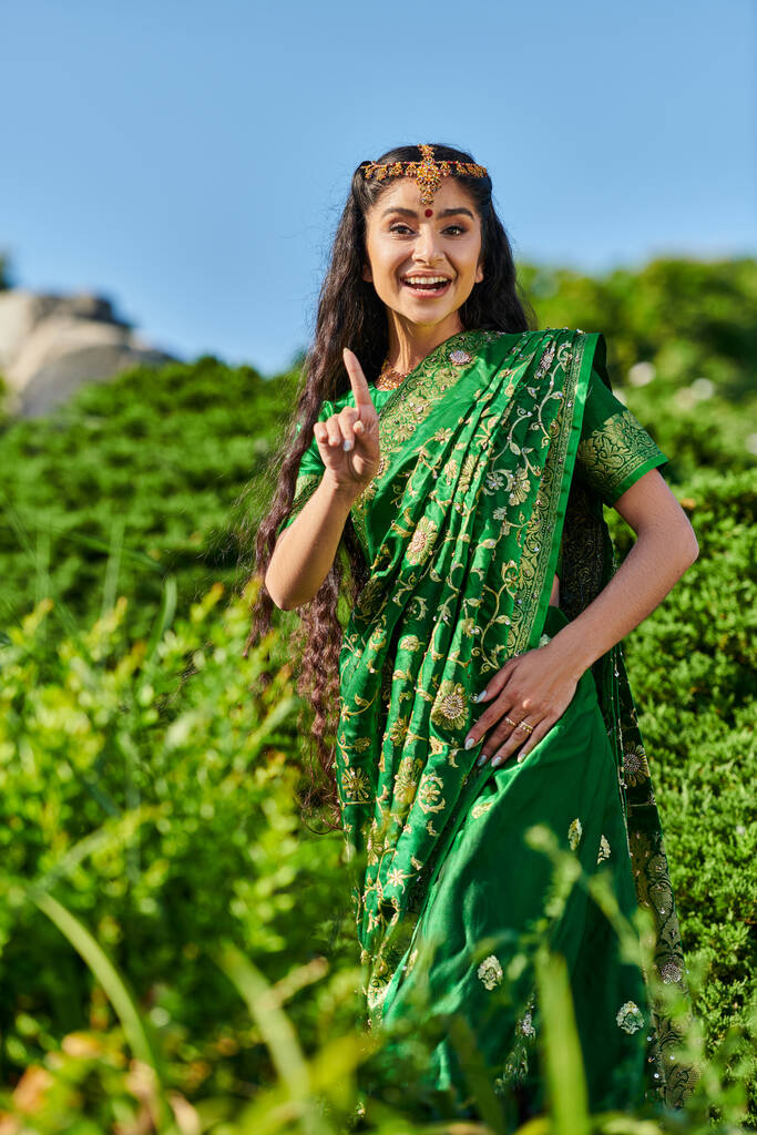 veselý mladý indián žena v sari ukazuje prstem, zatímco stojí v blízkosti rostlin v parku - Fotografie, Obrázek