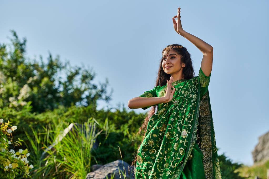 smiling indian woman in stylish green sari posing while standing in park with sky on background - Photo, Image