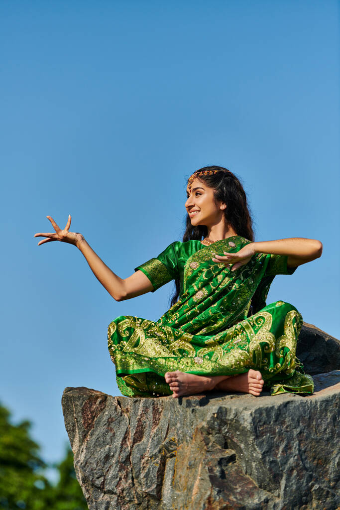 весёлая и стильная индийская женщина в зелёном сари позирует на камне с голубым небом на фоне - Фото, изображение