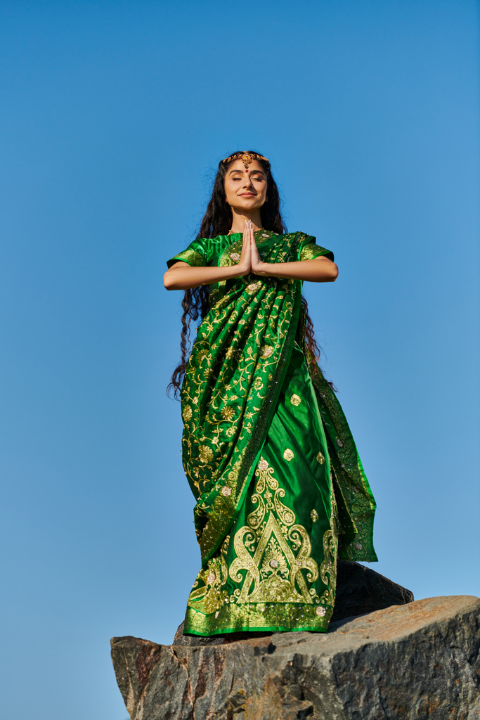 junge Indianerin in stylischem Sari meditiert auf Stein mit blauem Himmel auf Hintergrund - Foto, Bild