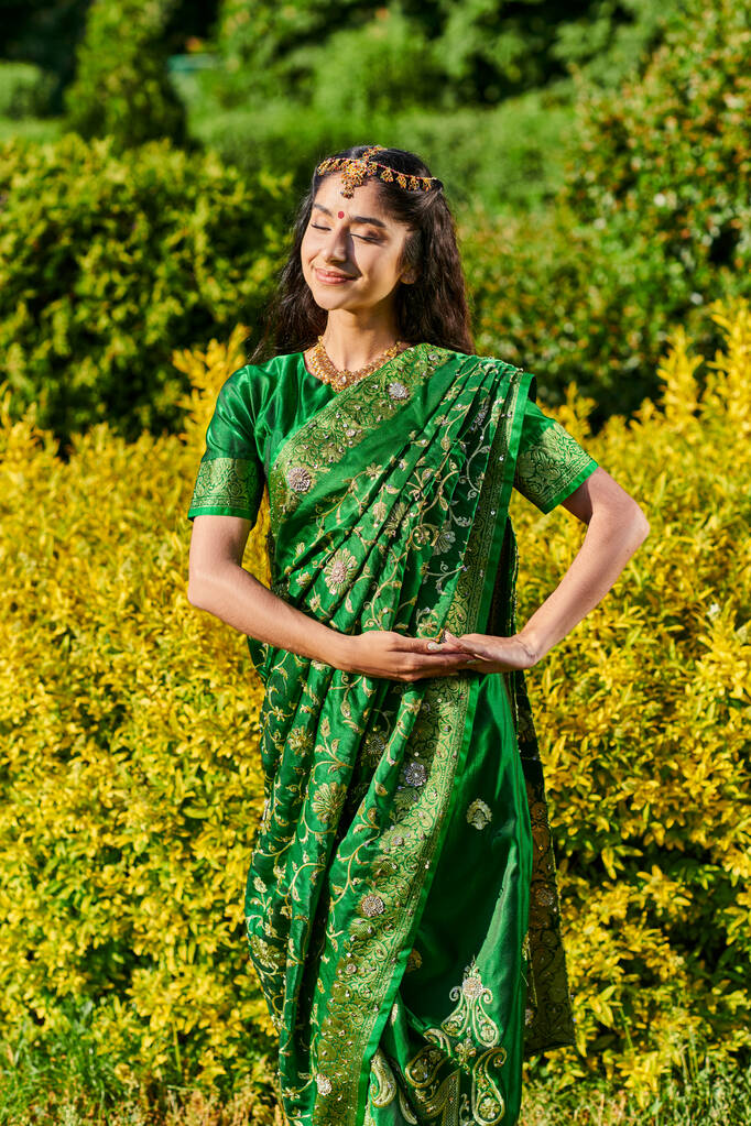 mujer india joven elegante y sonriente en sari posando en arbustos en el parque al aire libre - Foto, Imagen