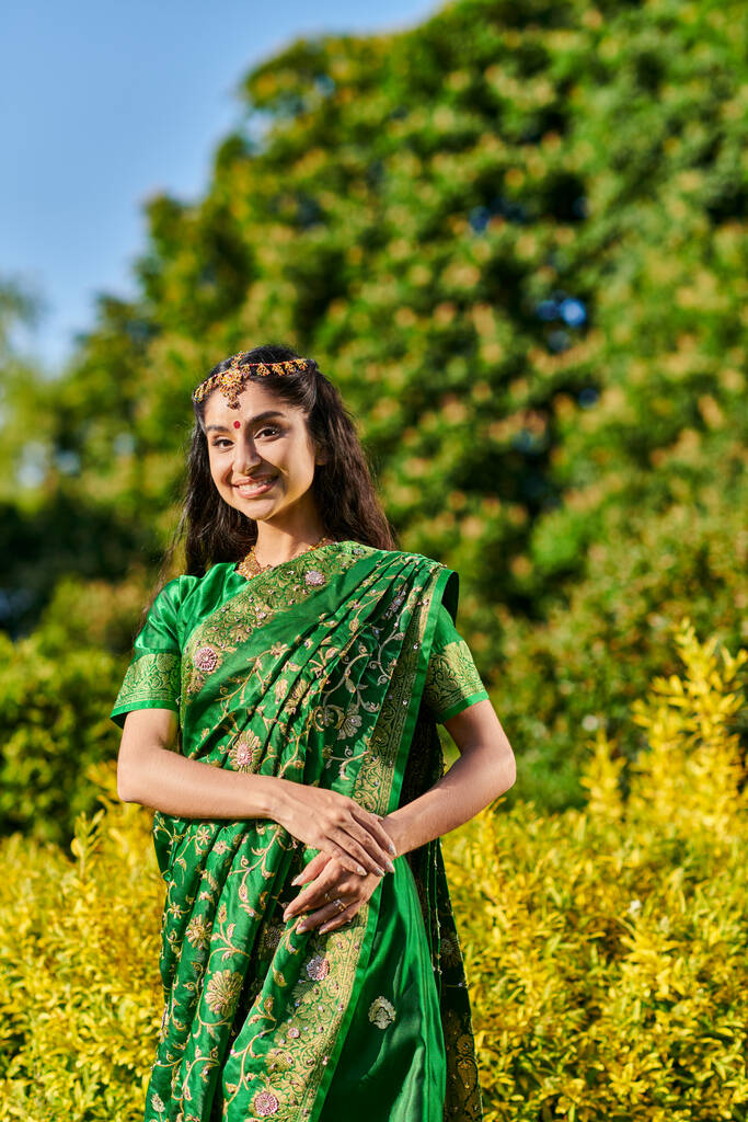 hübsche und lächelnde Indianerin im Sari, die in die Kamera schaut, während sie in der Nähe von Pflanzen im Park posiert - Foto, Bild