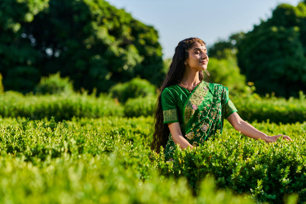 pleased young indian woman in sari standing with closed eyes near green plants in park - Photo, Image