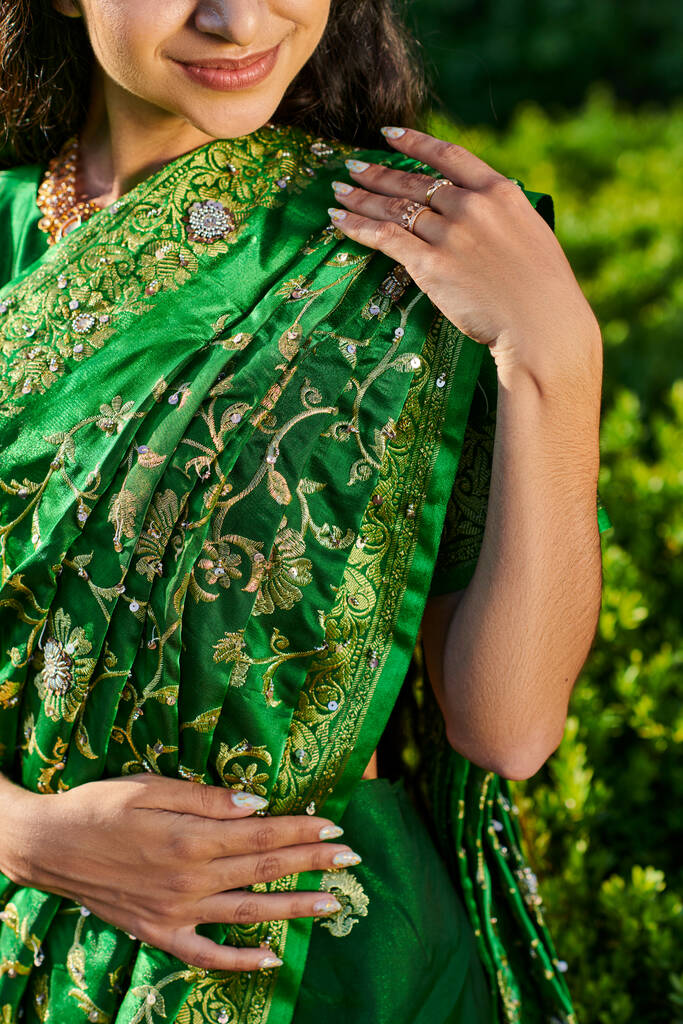 vista parziale di giovane donna sorridente ed elegante in moderno sari in piedi vicino a piante sfocate all'aperto - Foto, immagini