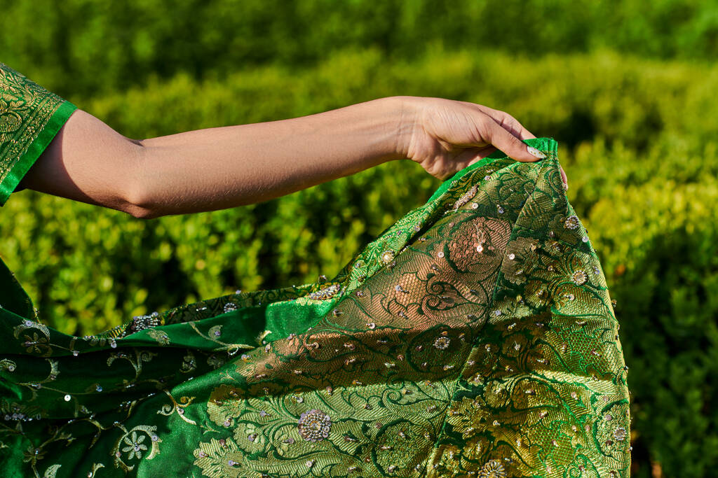 обрезанный вид молодой женщины, касающейся современного зеленого сари с узором рядом с растениями в парке - Фото, изображение