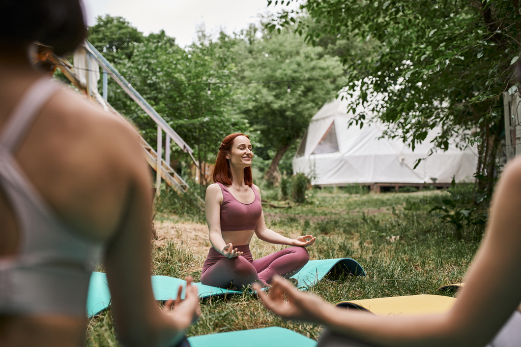 Unbekümmerte Frau meditiert in Lotus-Pose neben verschwommenen Freundinnen im Outdoor-Retreat-Zentrum - Foto, Bild