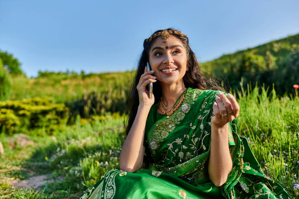 весела молода індійська жінка в модному сарі розмовляє по смартфону, сидячи на траві влітку - Фото, зображення