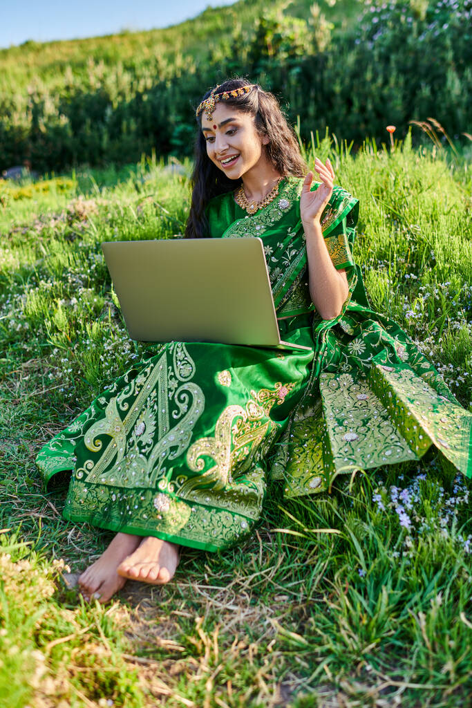 mujer india con estilo en sari tener videollamada en el ordenador portátil mientras está sentado en el prado en verano - Foto, imagen