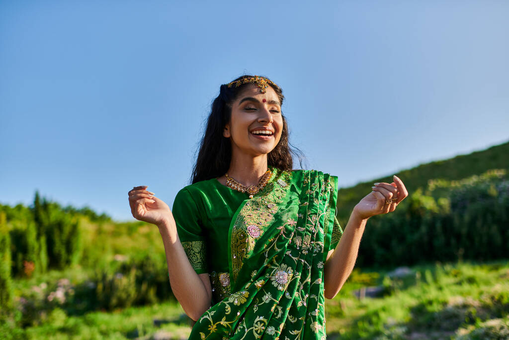 feliz jovem indiana em sari elegante posando e de pé com paisagem no fundo - Foto, Imagem
