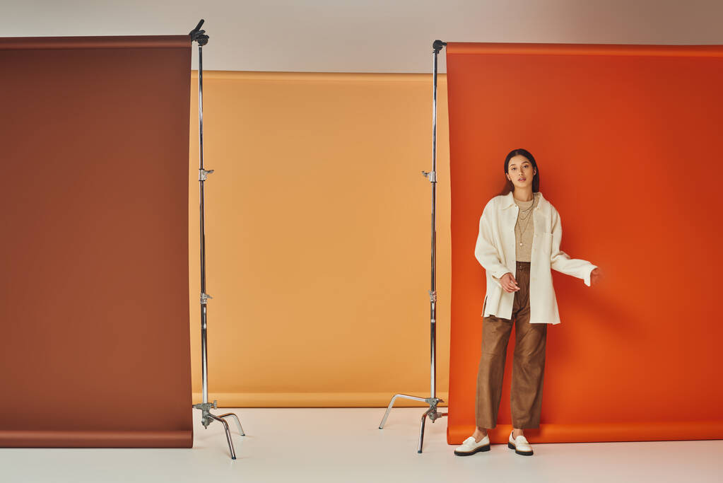 стильная азиатская модель в осеннем наряде, стоящая на разноцветном фоне, кожаных брюках и верхней одежде - Фото, изображение