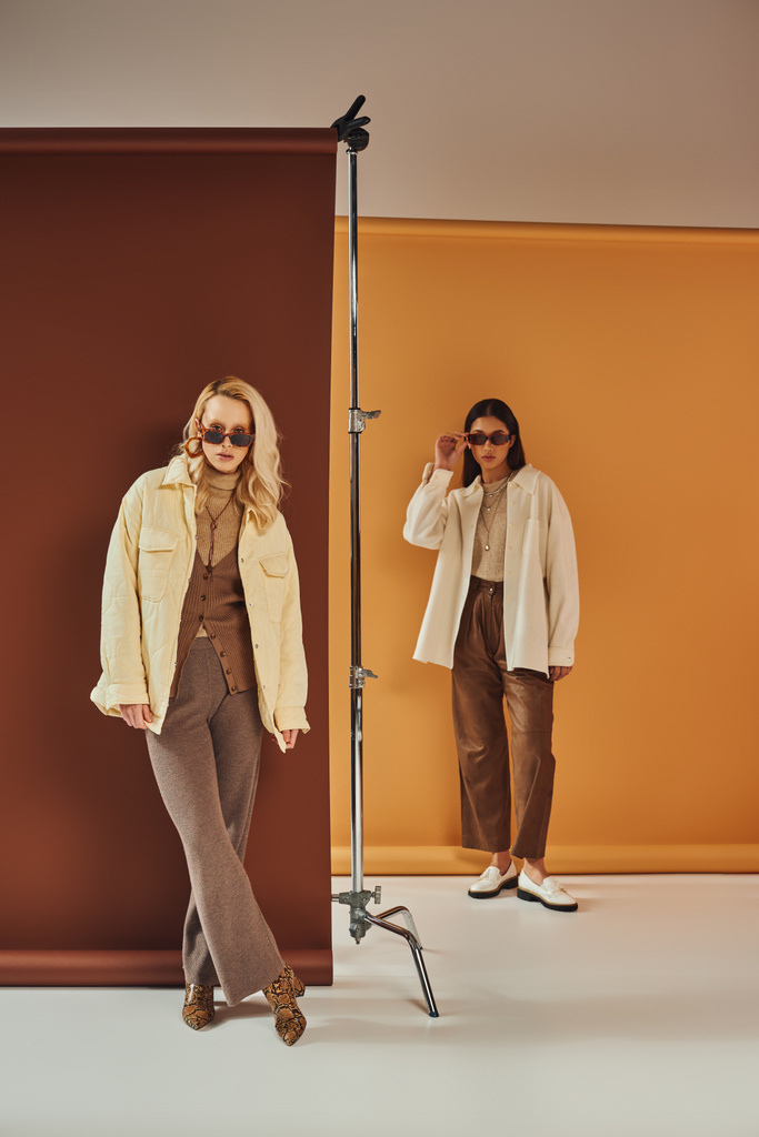 осенний сезон, мультиэтнические женщины в солнцезащитных очках и осенней верхней одежде позируют на фоне цвета дуэта - Фото, изображение