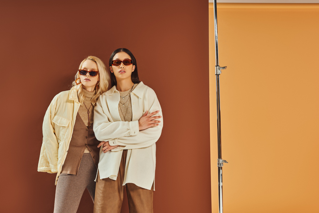 Осенняя мода и тенденции, межрасовые женщины в солнцезащитных очках и верхней одежде позируют на фоне цвета дуэта - Фото, изображение
