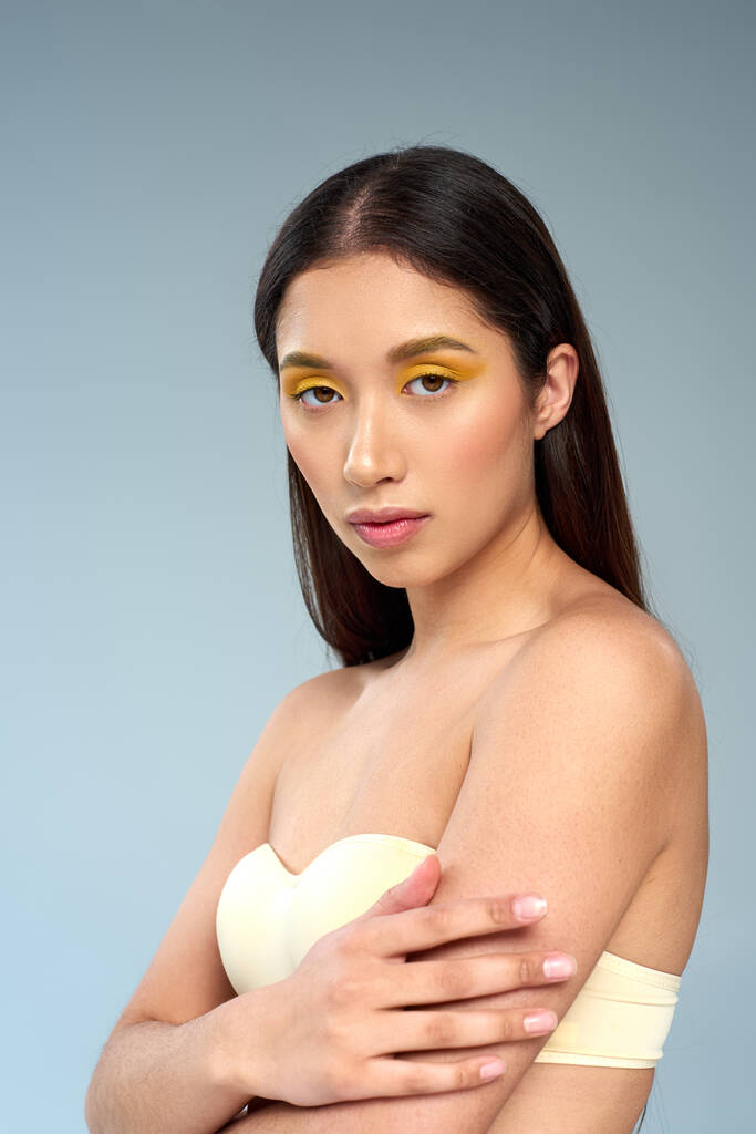 schoonheid en gratie, aziatisch model met gedurfde make-up en blote schouders poseren op een blauwe achtergrond - Foto, afbeelding