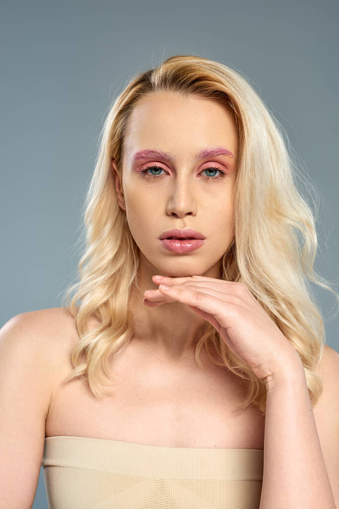 młoda kobieta z blond włosami i odważnym makijażem oczu patrząc na aparat fotograficzny na szarym tle, kobiece piękno - Zdjęcie, obraz