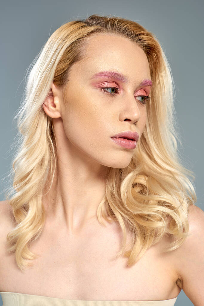 mujer sensual con maquillaje de ojos rosados y cabello rubio mirando hacia otro lado sobre fondo gris, belleza femenina - Foto, Imagen