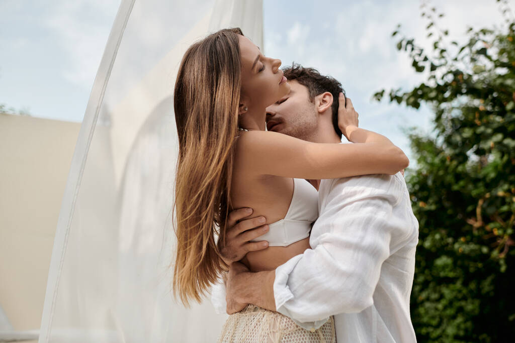 leidenschaftliches Paar, Mann küsst Frau den Hals, während er in der Nähe von weißem Tüll des Pavillons am Strand steht - Foto, Bild