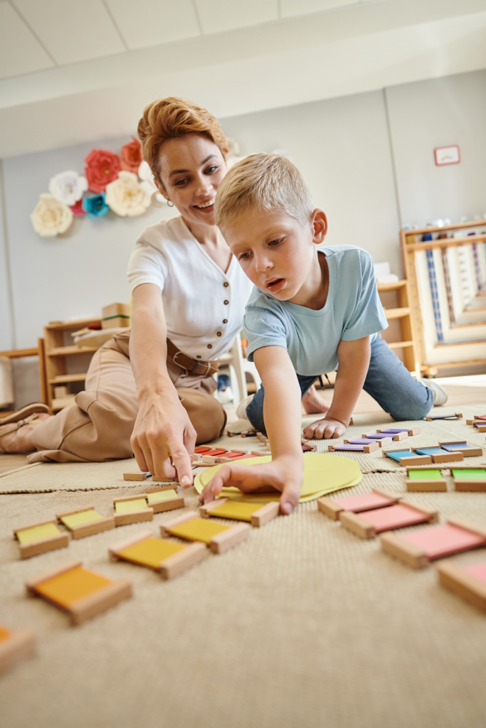 école montessori, garçon blond jouant jeu éducatif près de professeur femelle, mouvement, correspondance des couleurs - Photo, image