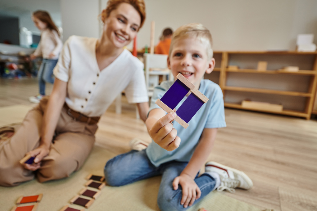 montessori σχολείο, χαρούμενο αγόρι παίζει το παιχνίδι ταιριάζουν χρώμα κοντά στο θηλυκό δάσκαλο, κάθεται στο πάτωμα - Φωτογραφία, εικόνα