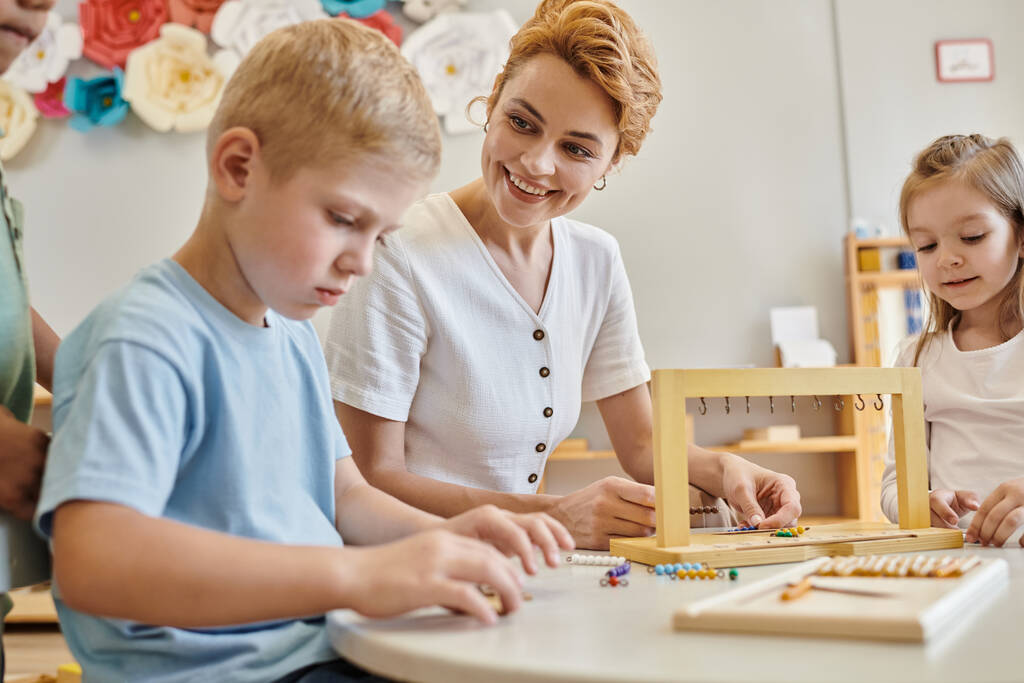 Montessori-Materialien, glückliche Lehrerin beobachtet konzentrierten Jungen in der Nähe von Kindern während des Unterrichts - Foto, Bild