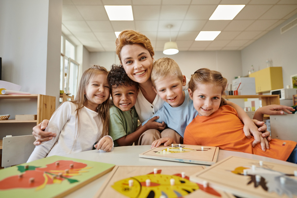 professor sorridente abraçando crianças multiétnicas perto de materiais didáticos na mesa na escola montessori - Foto, Imagem