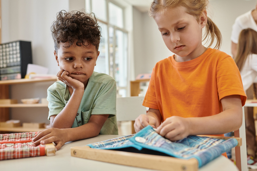 Afrykański amerykański chłopiec patrząc na przyjaciela bawiącego się tkaniną i guzikami w szkole Montessori - Zdjęcie, obraz