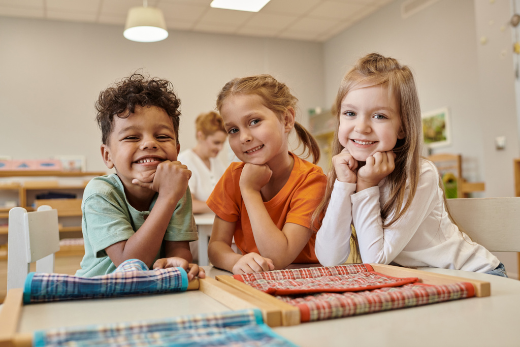 веселые и многонациональные дети, смотрящие в камеру возле стола в классе Монтессори - Фото, изображение