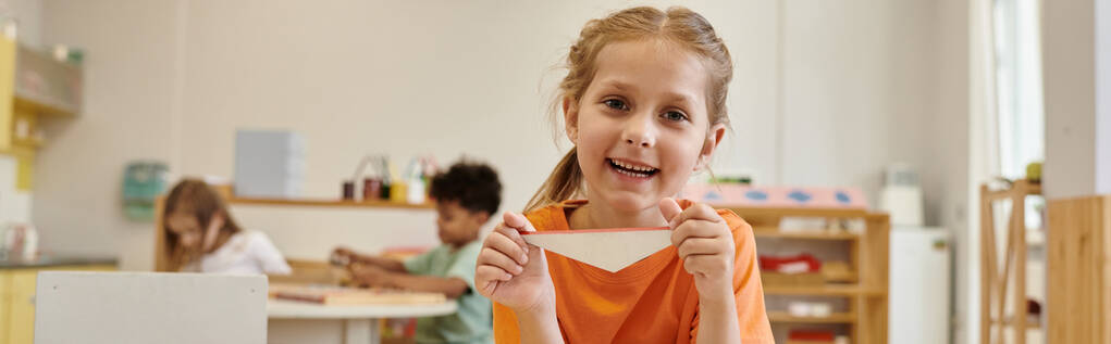 веселая девушка смотрит в камеру и держит треугольник в школе Монтессори, баннер - Фото, изображение