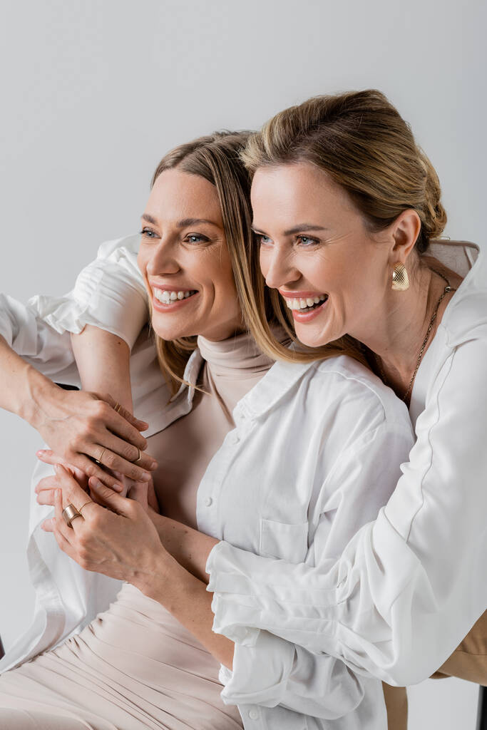 twee vrolijke voguish zussen in formele dragen knuffelen glimlachen en weg te kijken, stijl en mode - Foto, afbeelding