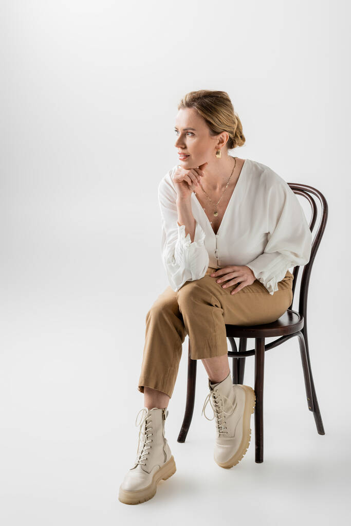 Blonde Frau in pastellfarbener Kleidung sitzt auf Stuhl und schaut weg, Hand an Kinn, Stil und Mode - Foto, Bild