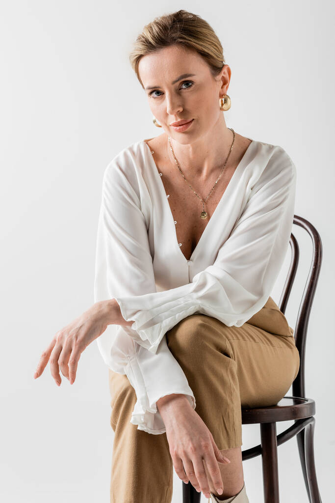 mujer rubia elegante sentada en la silla y relajándose con los brazos cruzados, estilo y moda - Foto, Imagen