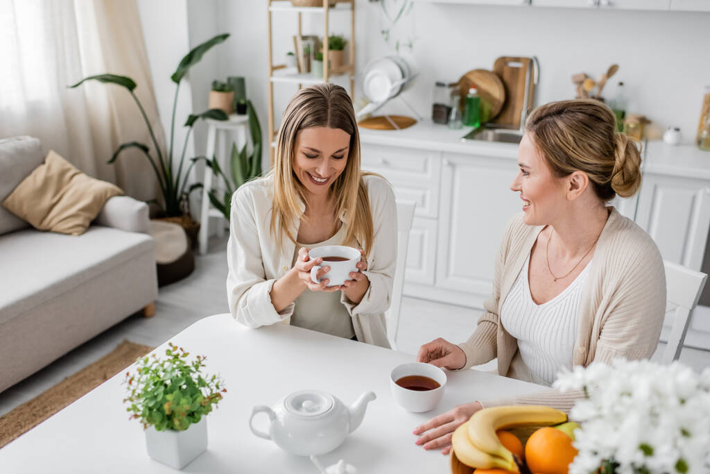 δύο ελκυστικές ευτυχισμένες αδελφές με περιστασιακή ενδυμασία κάθονται στο τραπέζι πίνοντας τσάι και χαμογελώντας, δένοντας - Φωτογραφία, εικόνα
