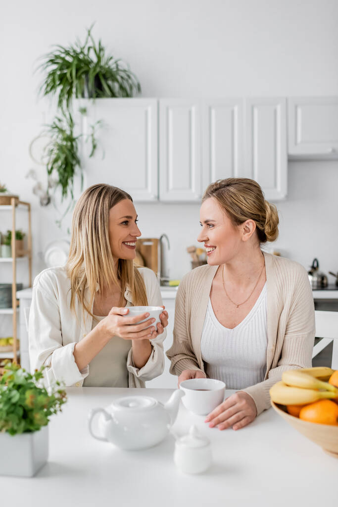 Закрыть две сестры, сидящие за столом, пьющие чай и улыбающиеся друг другу, семейные связи - Фото, изображение
