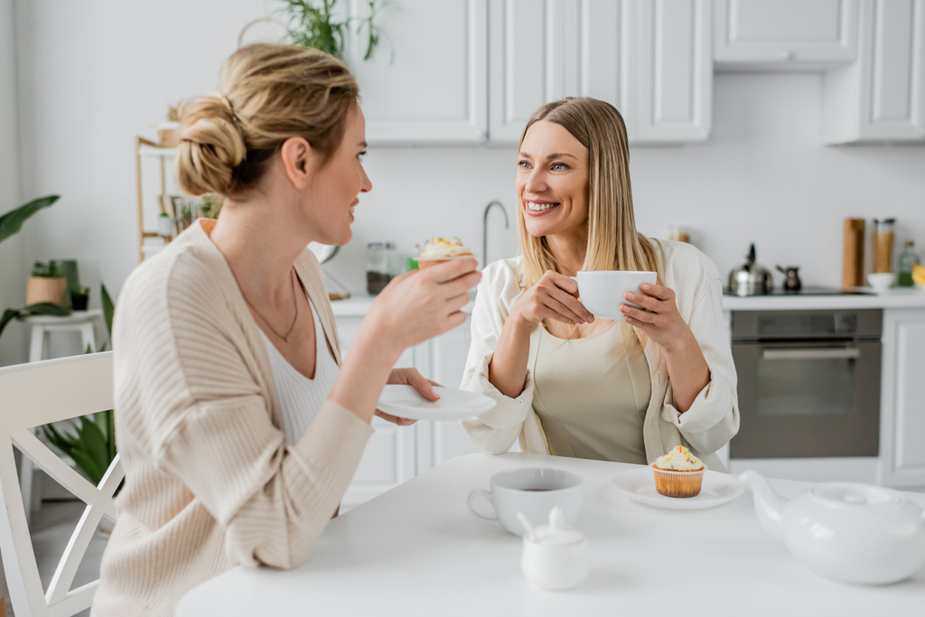 dobrze wyglądające siostry pijące herbatę i jedzące babeczki na tle mebli kuchennych, zacieśnianie więzi rodzinnych - Zdjęcie, obraz