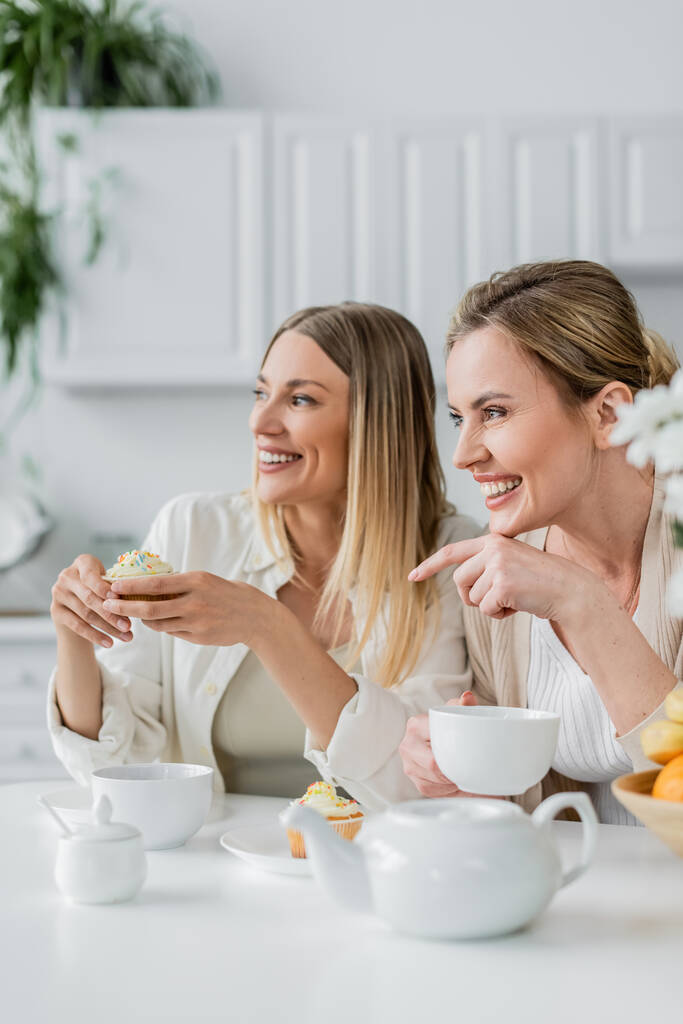 dos hermanas rubias guapas disfrutando de cupcakes y té y mirando hacia otro lado, señalando con el dedo, uniéndose - Foto, Imagen
