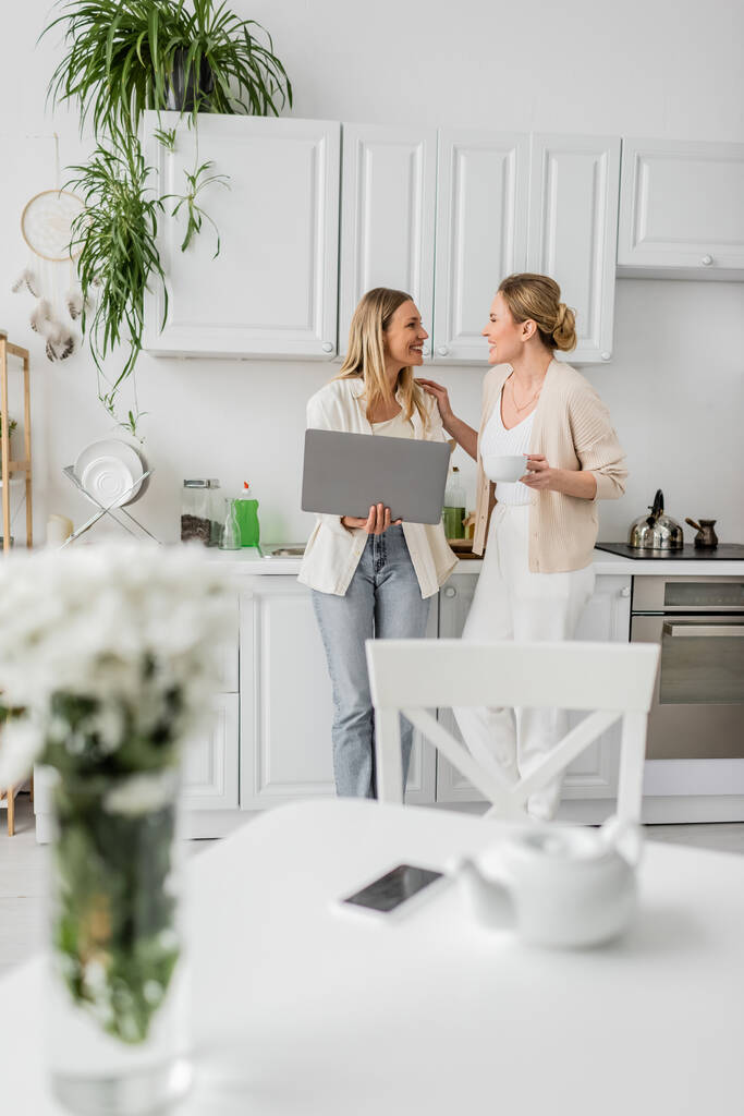 aantrekkelijke blonde zussen staan in de keuken met laptop kijken naar elkaar, familie binding - Foto, afbeelding