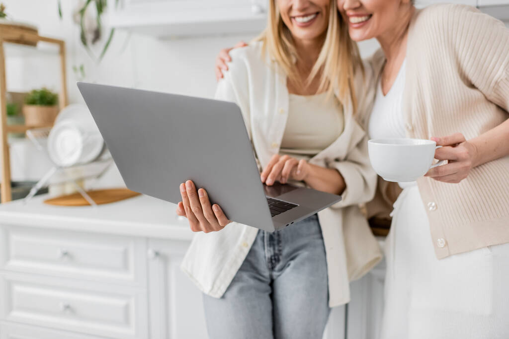 abgeschnittene Ansicht von zwei Smiley-blonden Schwestern in trendigen pastellfarbenen Strickjacken, die auf Laptop schauen, - Foto, Bild