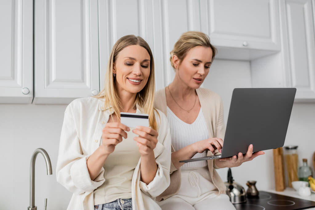 bom olhar irmãs encantadoras na moda traje pastel segurando cartão de crédito e laptop, ligação familiar - Foto, Imagem