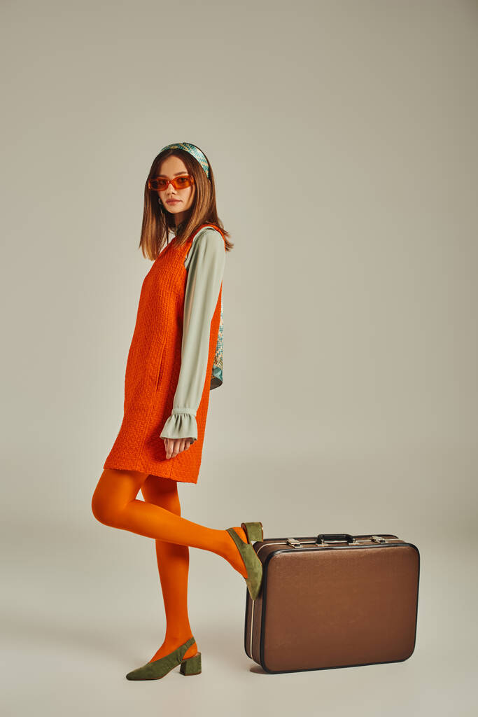 donna alla moda in abito arancione e occhiali da sole in posa vicino a valigia vintage su grigio, stile retrò - Foto, immagini