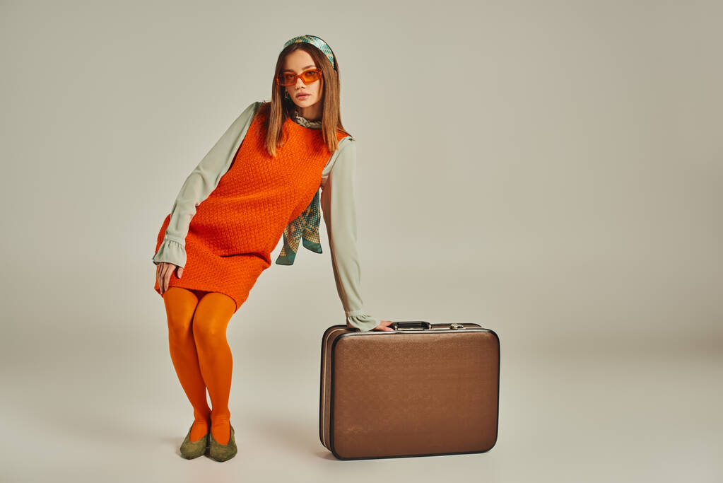 volledige lengte van glamour vrouw in oranje jurk poseren in de buurt vintage koffer op grijs, mode uit het verleden - Foto, afbeelding
