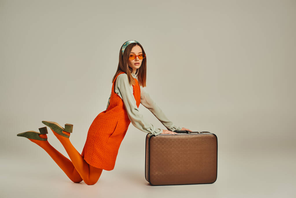 młoda kobieta w stylowych ubraniach retro i okularach przeciwsłonecznych klęcząc przy zabytkowej walizce na szarości - Zdjęcie, obraz