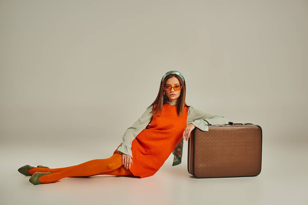 modelo joven en traje vintage de moda y gafas de sol sentados cerca de la maleta vintage en gris - Foto, imagen