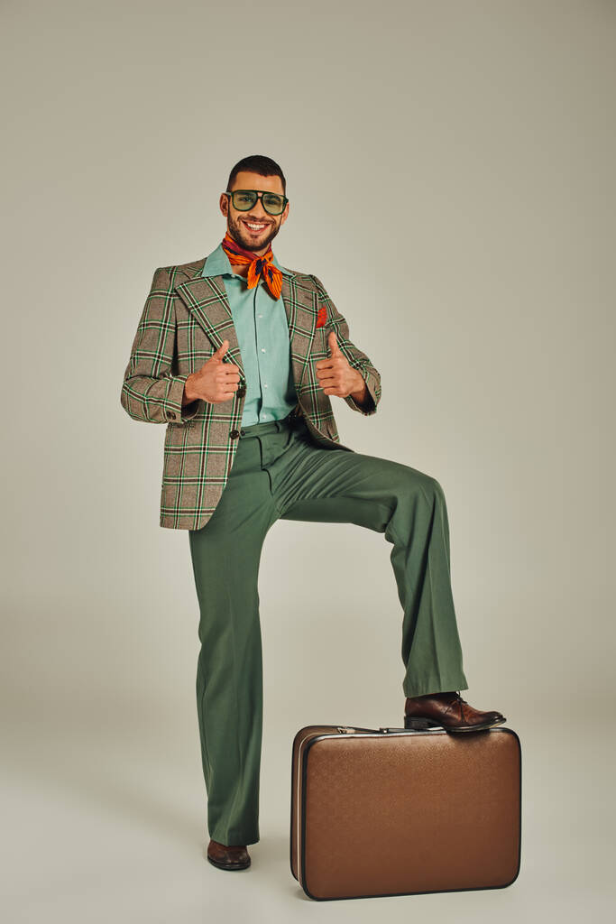 Eski moda giysiler ve güneş gözlüğü takan mutlu adam, gri bavulun yanında baş parmak gösteriyor. - Fotoğraf, Görsel