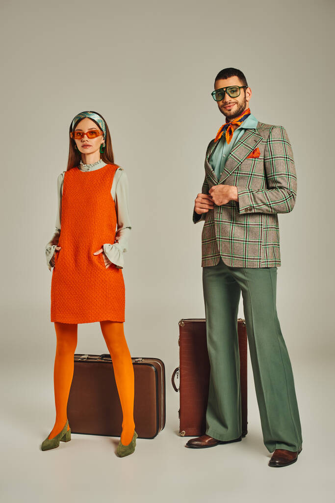 Frau in orangefarbenem Kleid und Sonnenbrille neben glücklichem Mann im karierten Blazer und Vintage-Koffer auf grau - Foto, Bild
