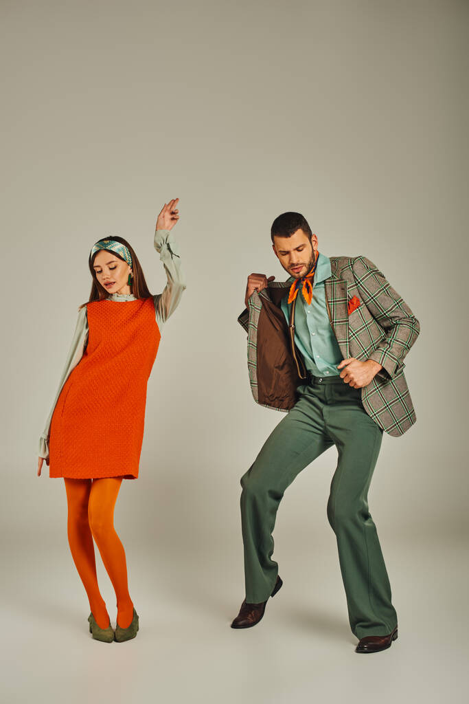 młoda para w marynarce w kratę i pomarańczowej sukience tańcząca na szarości, moda vintage, pełna długość - Zdjęcie, obraz