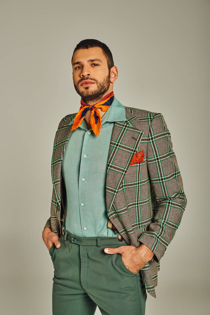 zelfverzekerde man in geruite blazer en kleurrijke halsdoek staand met handen in zakken op grijs - Foto, afbeelding
