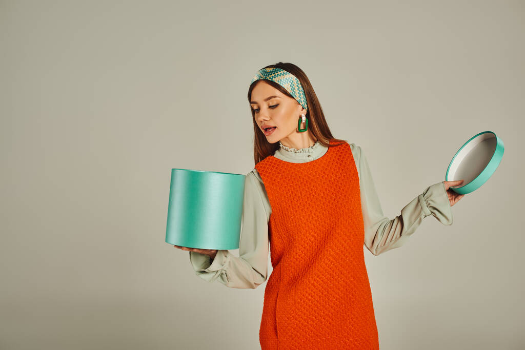 zvědavá žena v oranžových šatech a čelence hledající v dárkové krabici na šedé, retro módní fotografii - Fotografie, Obrázek