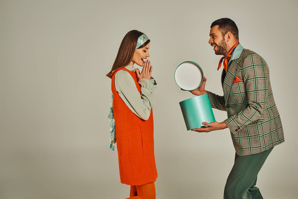 μοντέρνο άνδρα άνοιγμα κουτί δώρου κοντά έκπληξη γυναίκα σε πορτοκαλί φόρεμα σε γκρι, ρετρό εμπνευσμένο ζευγάρι - Φωτογραφία, εικόνα