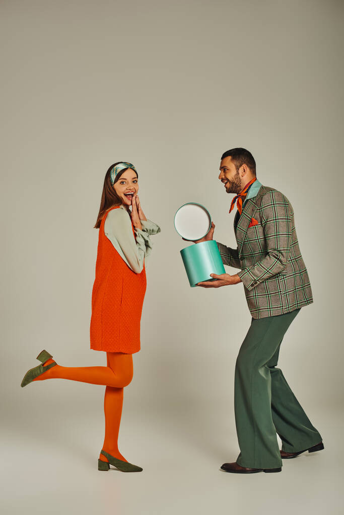 elegante hombre sosteniendo caja de regalo cerca de mujer llena de alegría en vestido naranja en gris, moda de inspiración retro - Foto, imagen