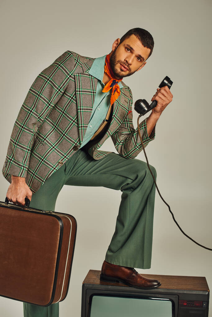 mężczyzna z walizką trzymając słuchawkę z telefonu komórkowego w pobliżu zabytkowego telewizora na szarym, retro stylu życia - Zdjęcie, obraz