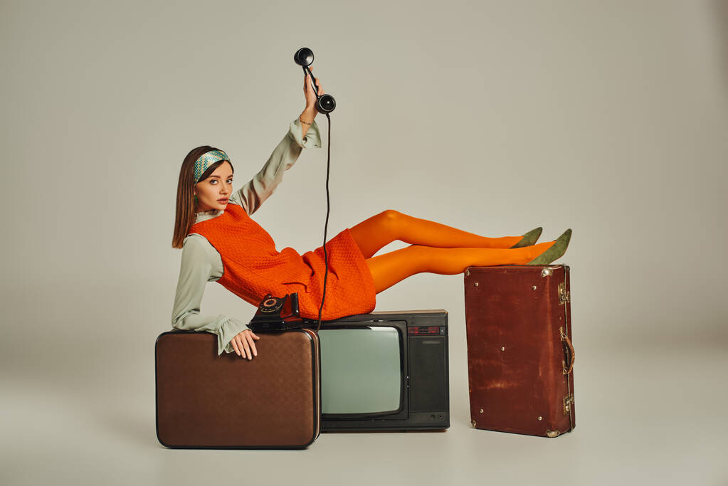 fiatal nő stílusos retro öltözékben ül vintage telefon TV-készülék és bőröndök szürke - Fotó, kép
